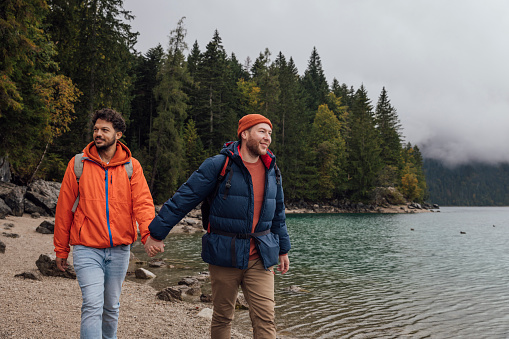 A same sex couple wearing waterproofs, exploring lake Eibsee while on holiday in Garmisch-Partenkirchen, Germany. They are walking by the water's edge on a pebble beach while holding hands and smiling.