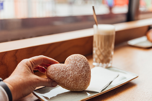 An unrecognisable mature woman sitting at a wooden bench in a cafe while on holiday in Garmisch-Partenkirchen, Germany. She is about to drink a latte and enjoy a heart shaped doughnut. She is holding it up towards the camera slightly.