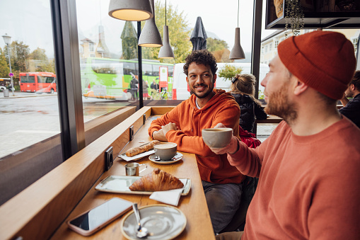 A same sex couple sitting in a cafe enjoying a baked pastry and coffee while on holiday in Garmisch-Partenkirchen, Germany. They are bonding and talking together while sitting at a high bench at the window and relax with a cup of coffee.