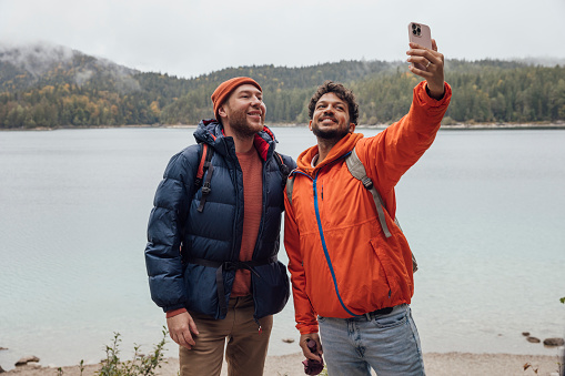 A same sex couple wearing waterproofs and backpacks exploring lake Eibsee while on holiday in Garmisch-Partenkirchen, Germany. They are taking a selfie together on a mobile phone with the lake in the background.