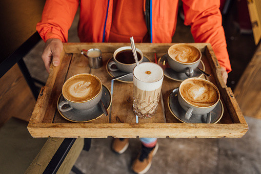 An unrecognisable mid adult man carrying a wooden tray with different coffees on it in a cafe while on holiday in Garmisch-Partenkirchen, Germany. He is taking over to a table with his friends on it for them to all enjoy.