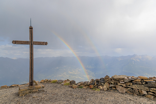 A scenics view of a mountain summit cross with a majestic double rainbow in the background under a stormy and rainy weather conditions