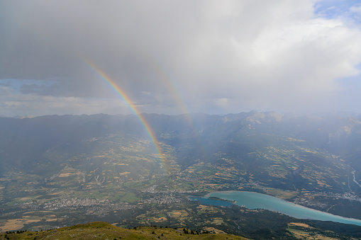 A scenics view of a mountain summit with a majestic double rainbow and Embrun, Hautes-alpes, France in the background under a stormy and rainy weather conditions