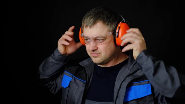 A man in work overalls puts on noise-canceling ear protectors, earmuffs on a black background.