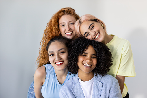A group of beautiful young women from different races, smiling and looking at the camera against a white wall backdrop. Concept of cross-cultural friendship and diverse beauty. Focus on African American girl.