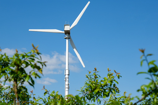 Green energy concept: Huge white wind turbine against a blue sky with bushes, twigs and branches with leaves in the blurred foreground, selective focus, copy space