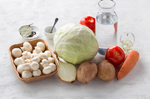 ngredients for vegetable stew: cabbage, mushrooms, potatoes, onions, carrots, tomatoes, salt, vegetable oil, spices and water on a light gray background. Cooking delicious vegan food.