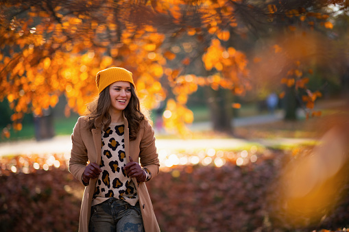 Hello november. smiling modern 40 years old woman in beige coat and orange hat outdoors in the city park in autumn.