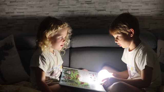 Two blond cute children, boy and girl, siblings, lying under the cover in bed, reading book together with small light, joy and happiness