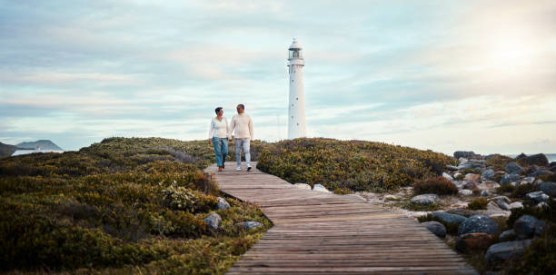 romance, amour et un couple se tenant la main en marchant sur la plage avec un phare en arrière-plan. nature, vue ou maquette de ciel bleu avec un homme et une femme faisant une promenade romantique à l’extérieur ensemble - senior adult outdoors wellbeing sky photos et images de collection