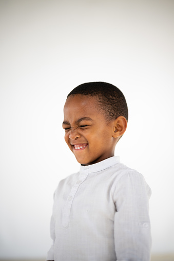 Portrait of a little boy of African ethnicity squeezing his eyes shut & pulling a funny face against a white background.