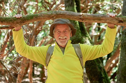 Happy bearded senior man hiking in the woods touching a moss covered tree trunk enjoying healthy lifestyle - earth day concept. Save the planet from deforestation