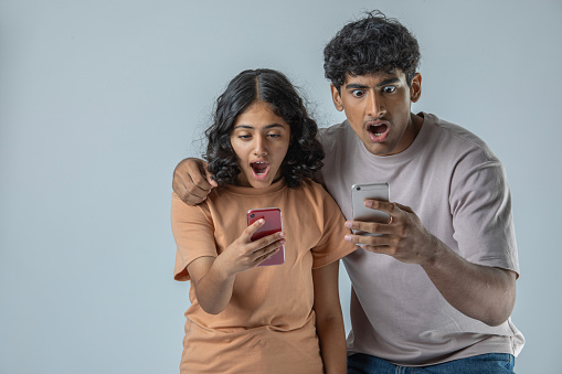 Shocked sister and brother with mouth open reading bad news over smart phones while standing against white background