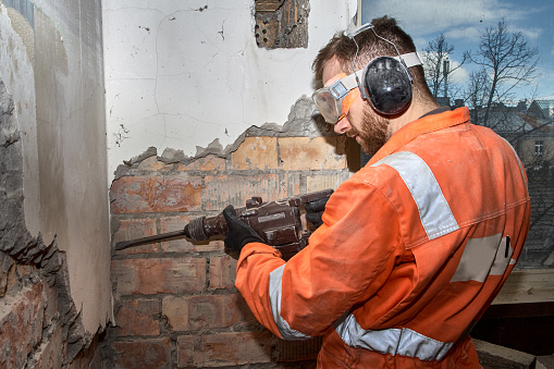 Construction worker using hammer drill with chisel for demolition jobs indoor, wearing orange coveralls, and PPE.