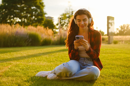 Happy girl wearing wireless headphones for listening to the music via smartphone in the park at sunset.