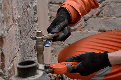 Plumber repairing leaking water connection on site, wearing gloves and orange coveralls.