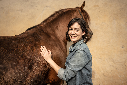 Portrait of a young woman petting her horse on a stable