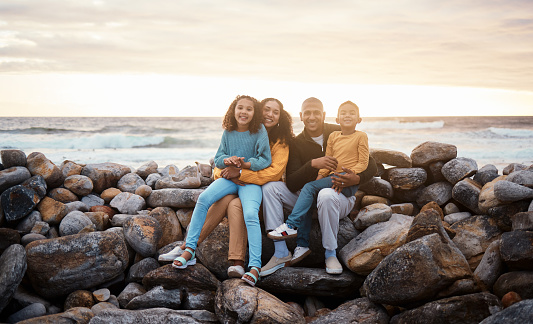 Black family, parents and children in beach portrait with excited face, sitting and rocks with happiness. Black woman, man and kids by ocean with love hug, care and bonding on holiday by sunset sky