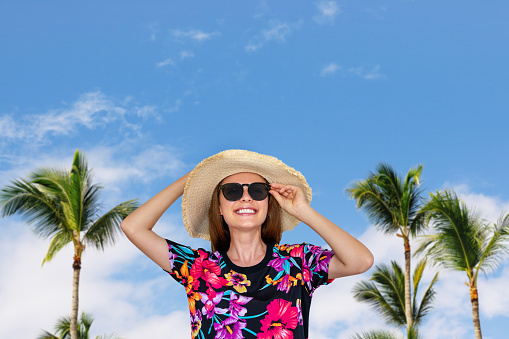 Woman wearing sun hat and sunglasses in front of palms tree and blue sky, travel background, summer vacation