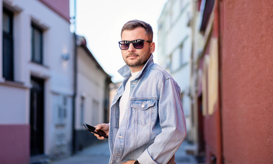 Trendy young adult man in sunglasses and denim jacket standing on street and holding mobile phone in his hand.