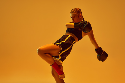 Dynamic photo of athletic teen girl, mma fighter in sportswear training against orange background in neon lights. Concept of mixed martial arts, sport, hobby, competition, athleticism, strength, ad