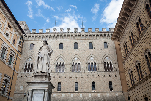 Siena, Italy - 2022, May 2 : The statue of  Sallustio Bandini, completed in 1880, standing in the Salimbeni square in front of the main building of the Monte dei Paschi di Siena bank