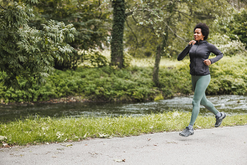Beautiful smiling mature African American woman is running along trail near the mountains river in nature.