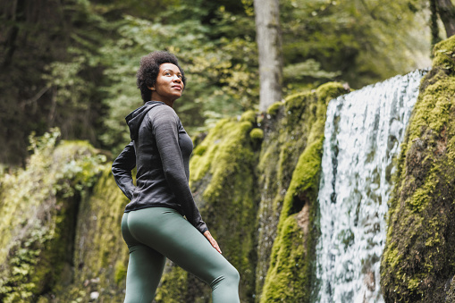 Mature African American female runner relaxing after jogging near the mountains waterfall in nature.