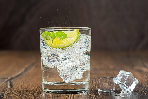 Cold, refreshing in the heat, sparkling water in a glass with ice cubes, lime wedge and mint leaves. Dark wooden bar counter background. Artificial acrylic ice.