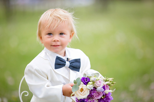 Beautiful toddler boy, dressed in white tuxedo, holding gorgeous flower bouquet for mothers day, smiling happily and giving it to mom in park