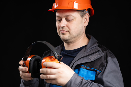 Builder in a protective helmet with noise-cancelling headphones in his hands on a black background.