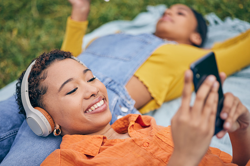 Music, phone and lesbian couple relax at picnic on grass, technology and streaming service app in nature. Cellphone, listening to headphones and happy lgbt women on blanket in garden together in park