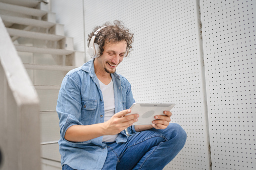 One caucasian man with curly hair outdoor in front of modern building use digital tablet and headphones to watch movie or series online stream or to have video call copy space