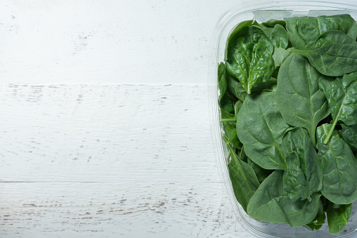 Clear, plastic container holding fresh spinach.  White, wooden background.