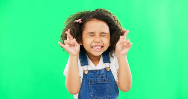 Photo of Fingers crossed, wish and praying with girl in studio for good luck, hope and optimism. Excited, emoji and closed eyes with child isolated on green background for miracle, positive and youth