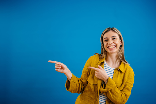 Portrait of a beautiful young woman pointing with her fingers while standing in front of a blue background.