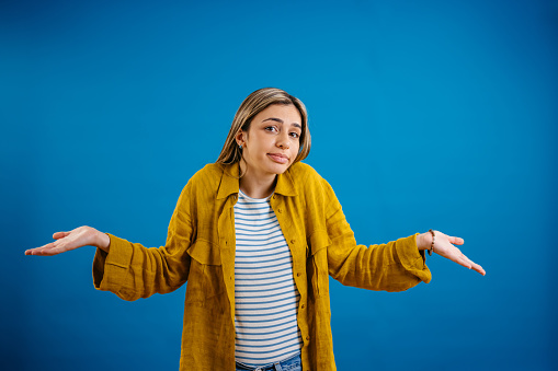 Portrait of a beautiful young woman shrugging her shoulders while standing in front of a blue background.