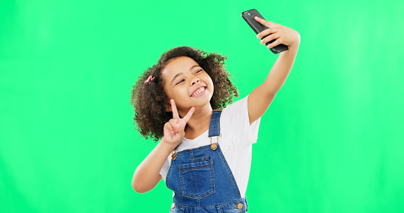 Phone, selfie and child peace sign in a studio with happiness from profile picture. Isolated, green background and young girl with technology showing a v hand gesture for social media with a smile