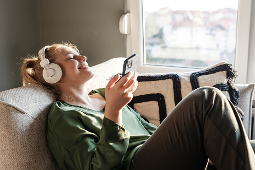 Young woman relaxing on a sofa and listening music on her phone via headphones