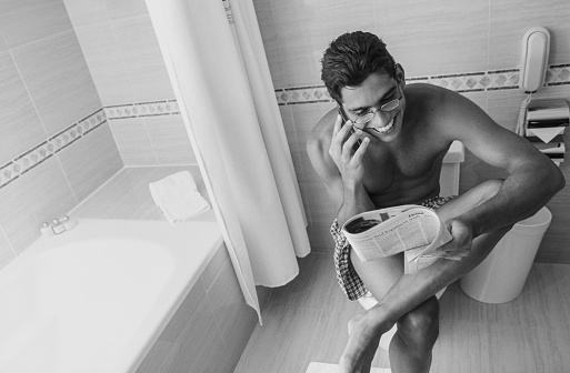 Man sitting in a hotel's bathroom using a mobile phone and reading a magazine