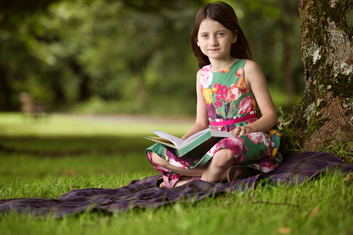 Primary age brunette, wearing a Summer dress, reading in the park.
