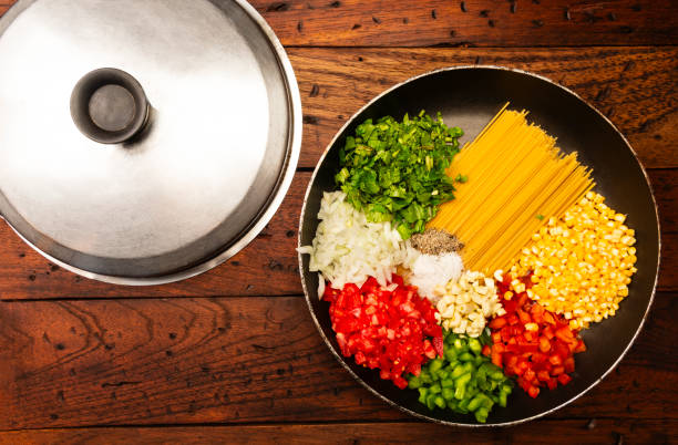 Uncooked pasta in a pan with the ingredients to be cooked . In the pan the ingredients are: raw spaghetti,chopped red pepper and green,chopped onion,chopped garlic,chopped tomato,sweet corn,salt,black peppercorn,cilantro. Image made in studio over rustic wood table.