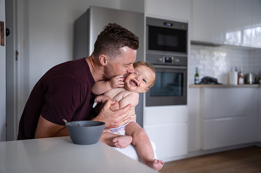 A loving, Caucasian father holds his 9 month old Eurasian son on his lap and gives him a kiss on the cheek as he spoon-feeds him a meal in the kitchen of their home.