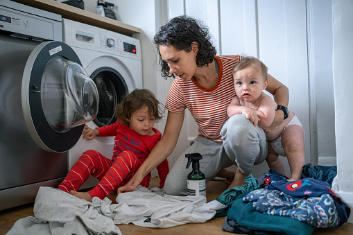 Eurasian mother holds her 9 month old son in her arms as she kneels on the ground of the laundry room, sorting dirty clothes with the help of her three year old daughter.