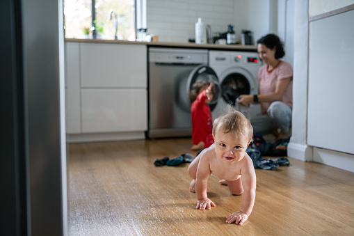 An adorable 9 month old Eurasian baby boy smiles and crawls towards the camera while in the background his mother changes out a load of laundry with the help of his 3 year old sister.