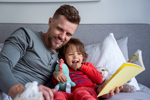 Adorable multiracial three year old girl of Hawaiian and Chinese descent lies in bed and laughs as her loving Caucasian dad reads a bedtime story to her.