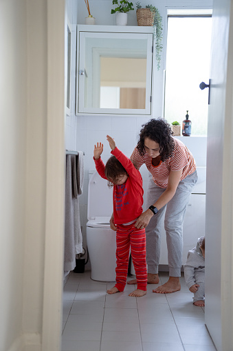 Three year old Eurasian girl stands with her arms up as her mother helps get her dressed in pajamas after potty training in the bathroom of their home.