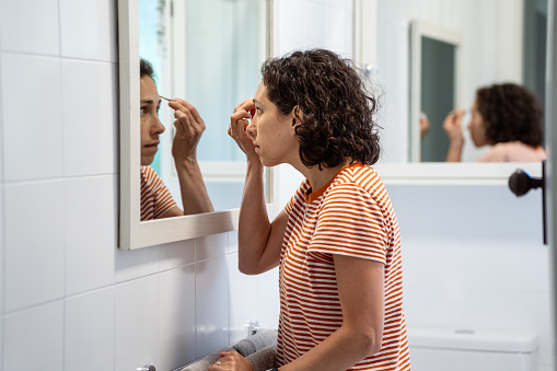 Young woman of Pacific Islander and Asian descent stands in front of the bathroom mirror and applies mascara as she gets ready in the morning.