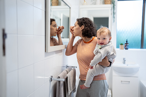 Eurasian mother holds her 9 month old son on her hip as she stands in front of a bathroom mirror and applies mascara as she gets ready for the day.