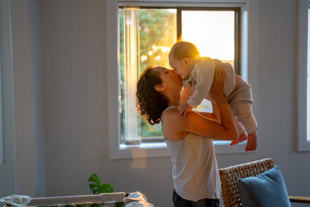 Loving mother holds her son up and kisses him in a bedroom Loving and affectionate Eurasian mother playfully lifts her 9 month old son above her head and kissing him on the cheek as they play together in a bedroom at sunset. baby boy dancing with his mommy stock pictures, royalty-free photos & images